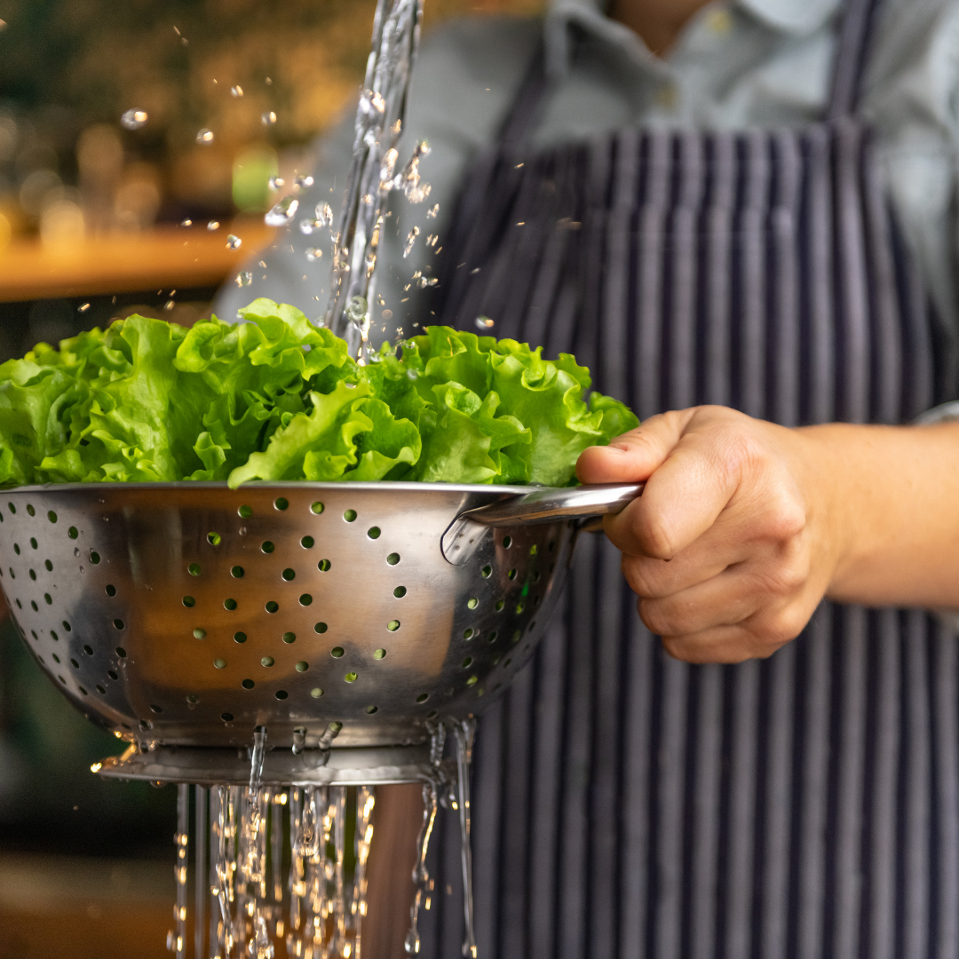 Person rinsing lettuce in a metal colander under running water.