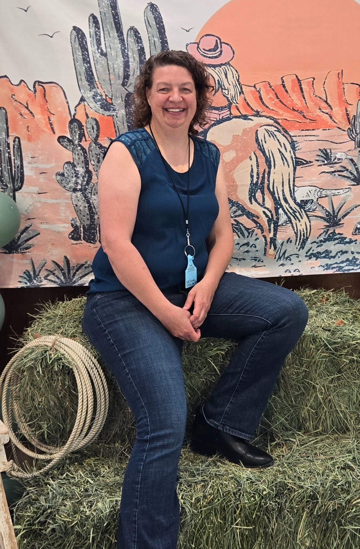 Amanda, who has curly brown hair, sits and smiles on a bale of hay with a painted cowboy background.
