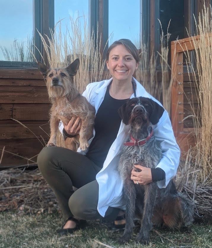 Chloe kneels on the ground and smiles, holding a smaller dog in her right hand and hugging another dog with her left hand.
