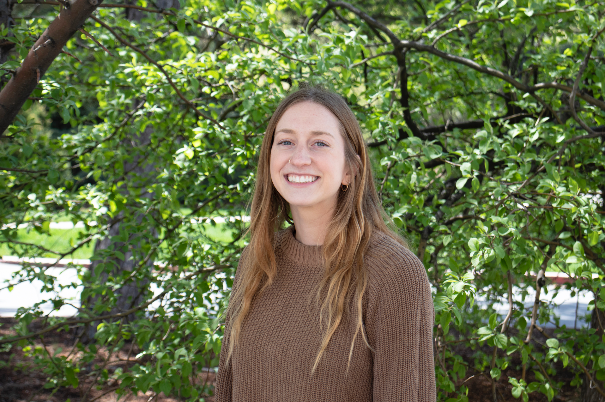 Emily, who has long dark blonde hair. smiles in front of trees.