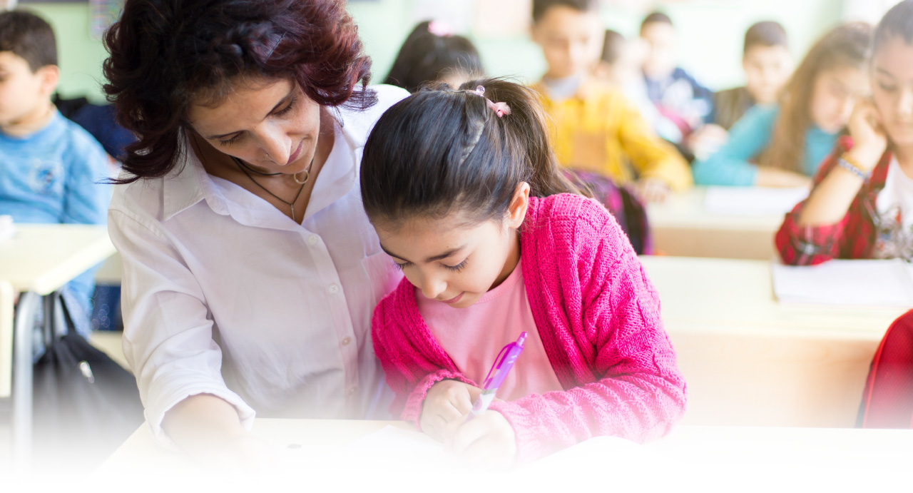 Teacher helps a young girl write in a classroom filled with other students.