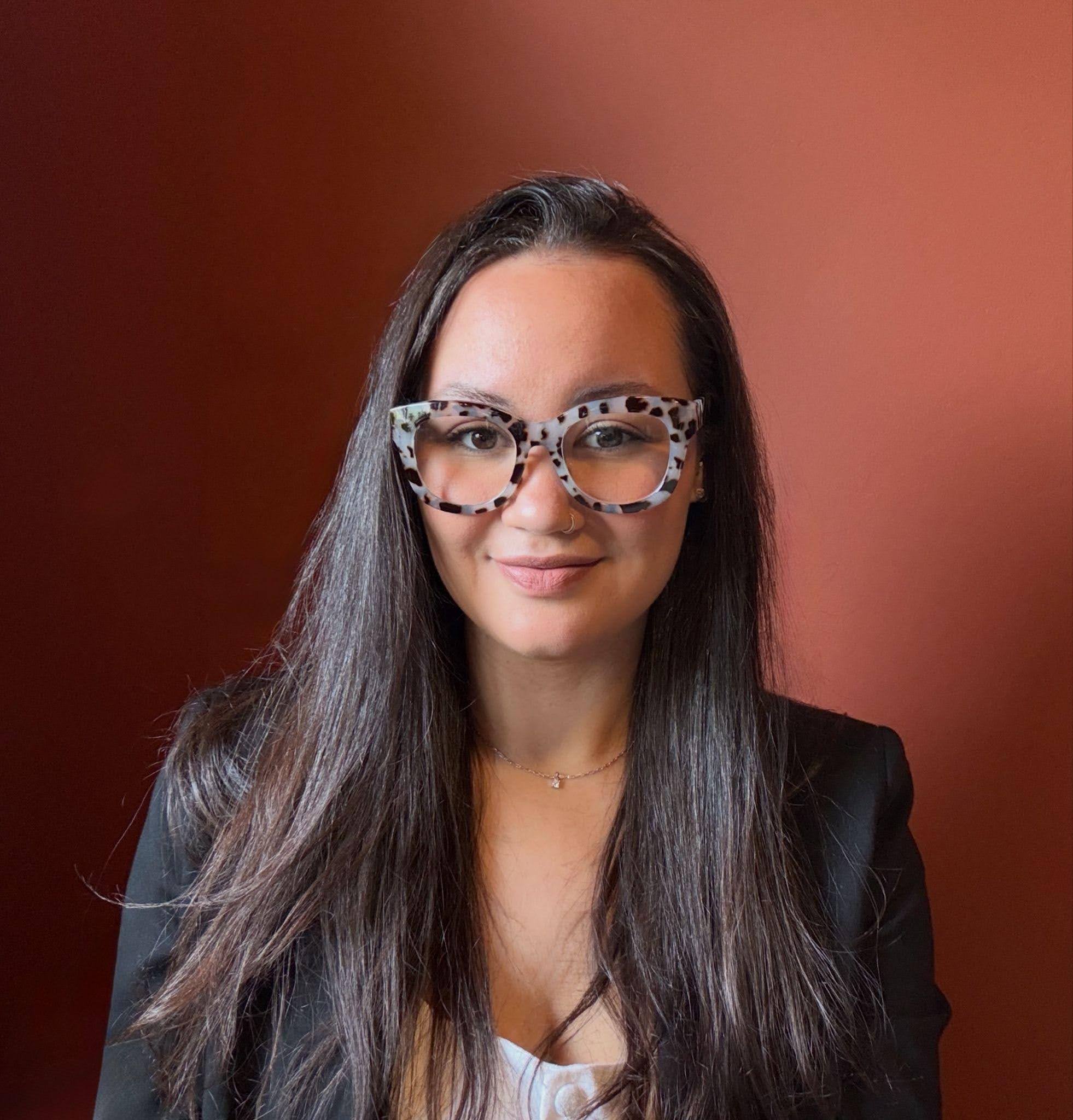 Jade, who has dark brown hair and cow-print classes, smiles in front of a dark orange background.