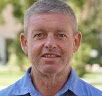 Korry, who has gray hair and is wearing a blue collared shirt, smiles at the camera.