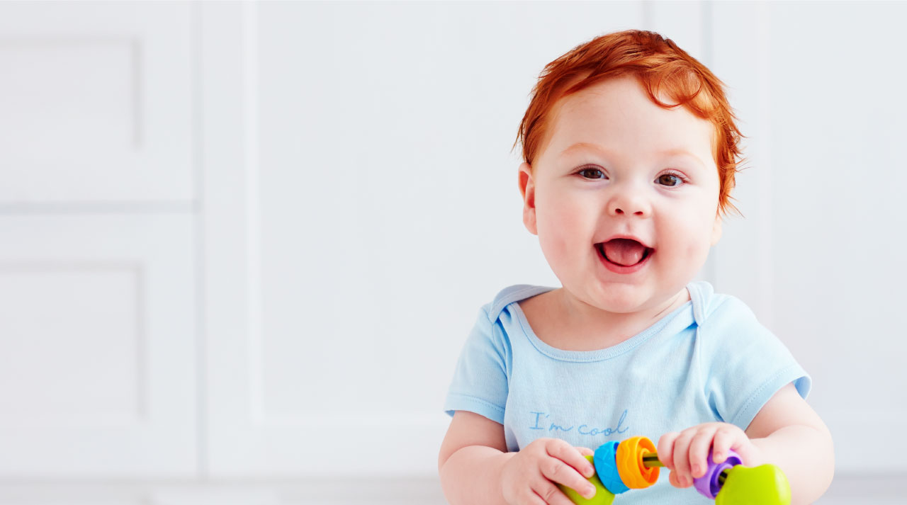 Smiling baby with red hair holds a colorful toy, wearing a light blue onesie indoors.