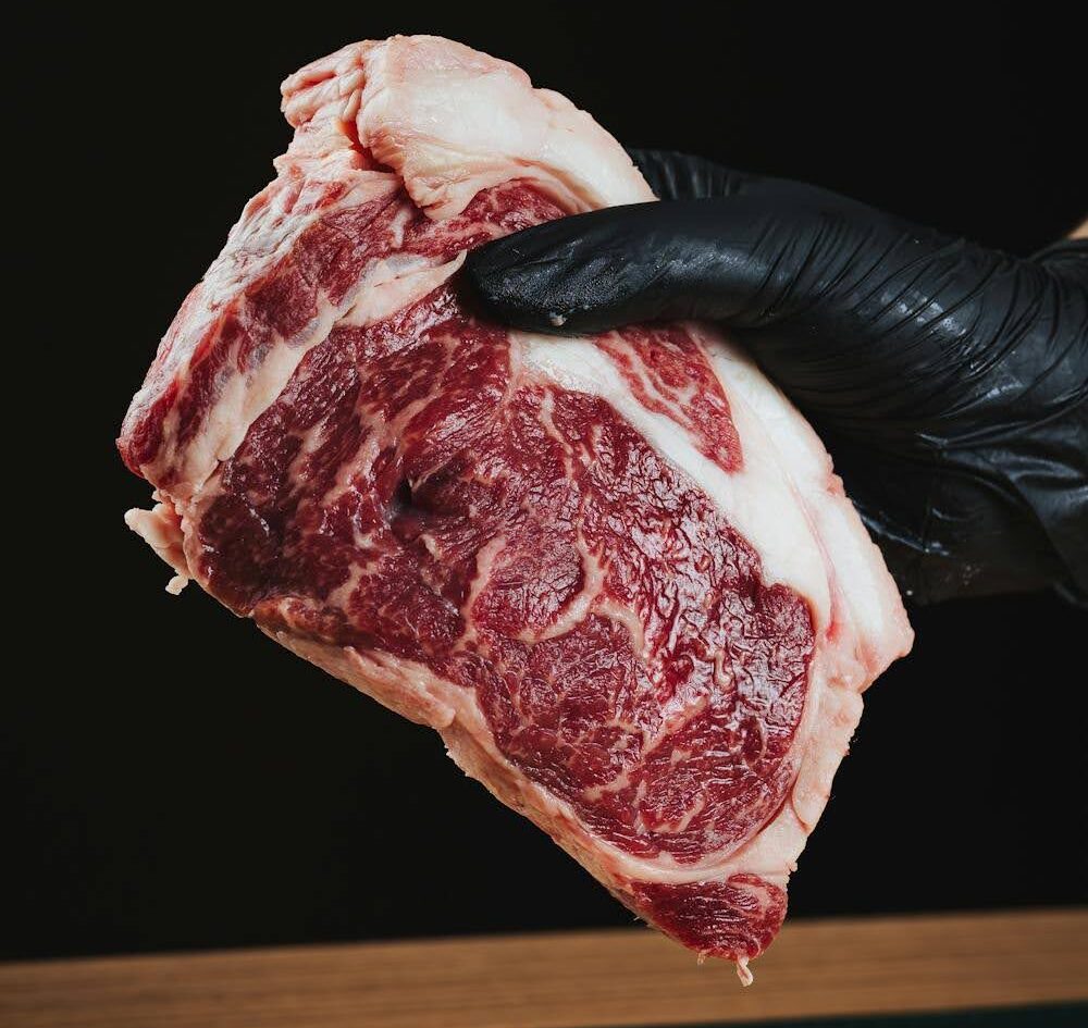 A gloved hand holds a raw marbled steak over a wooden surface with a black background.